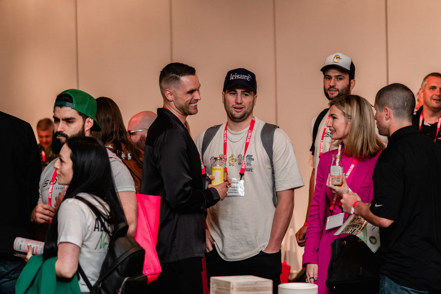 A group of men and women in lanyards stand talking, one in a navy Leisure cap and another in a green cap.