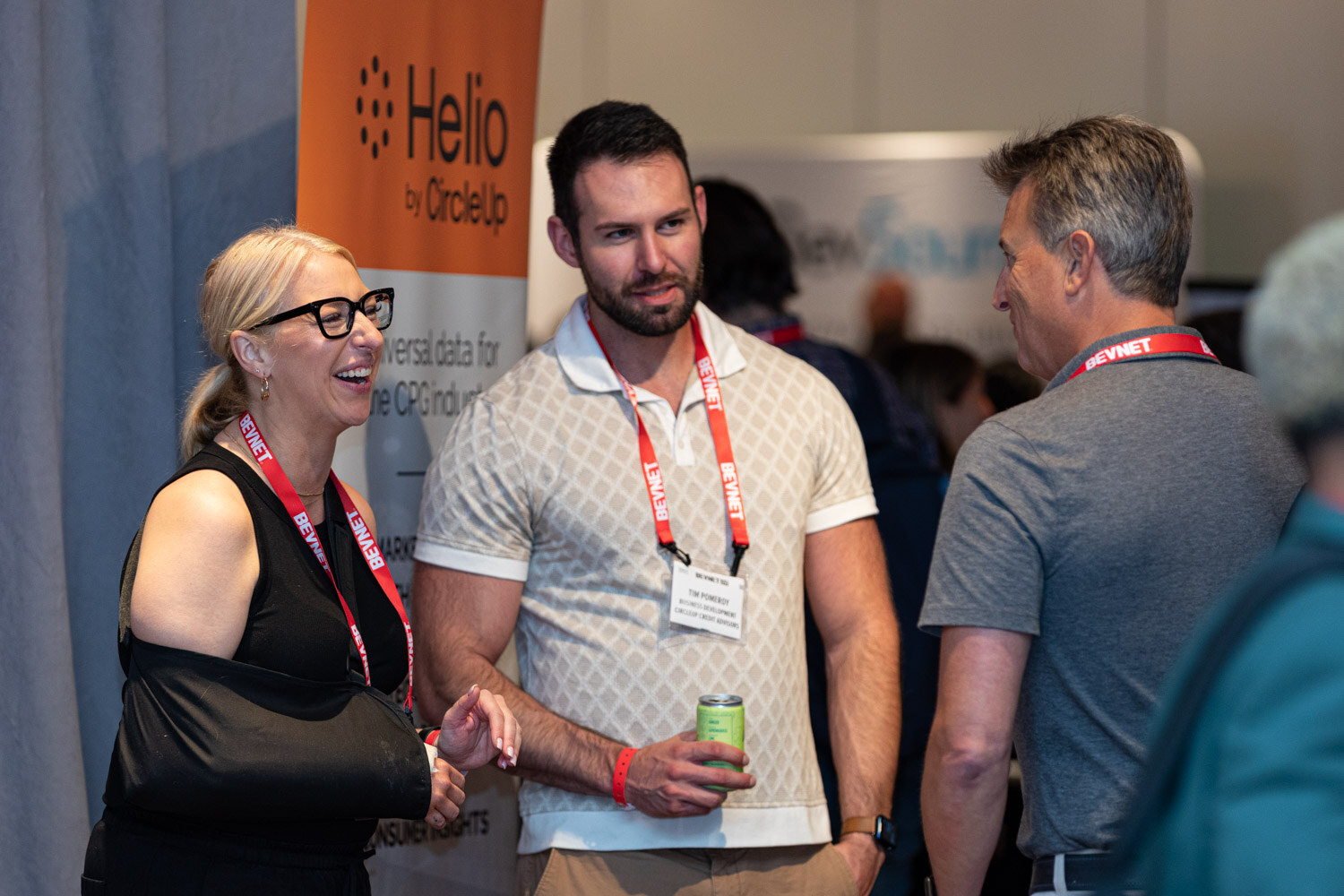 A woman with a black arm sling and glasses and two men in lanyards smile in front of an orange Helio by CircleUp banner.