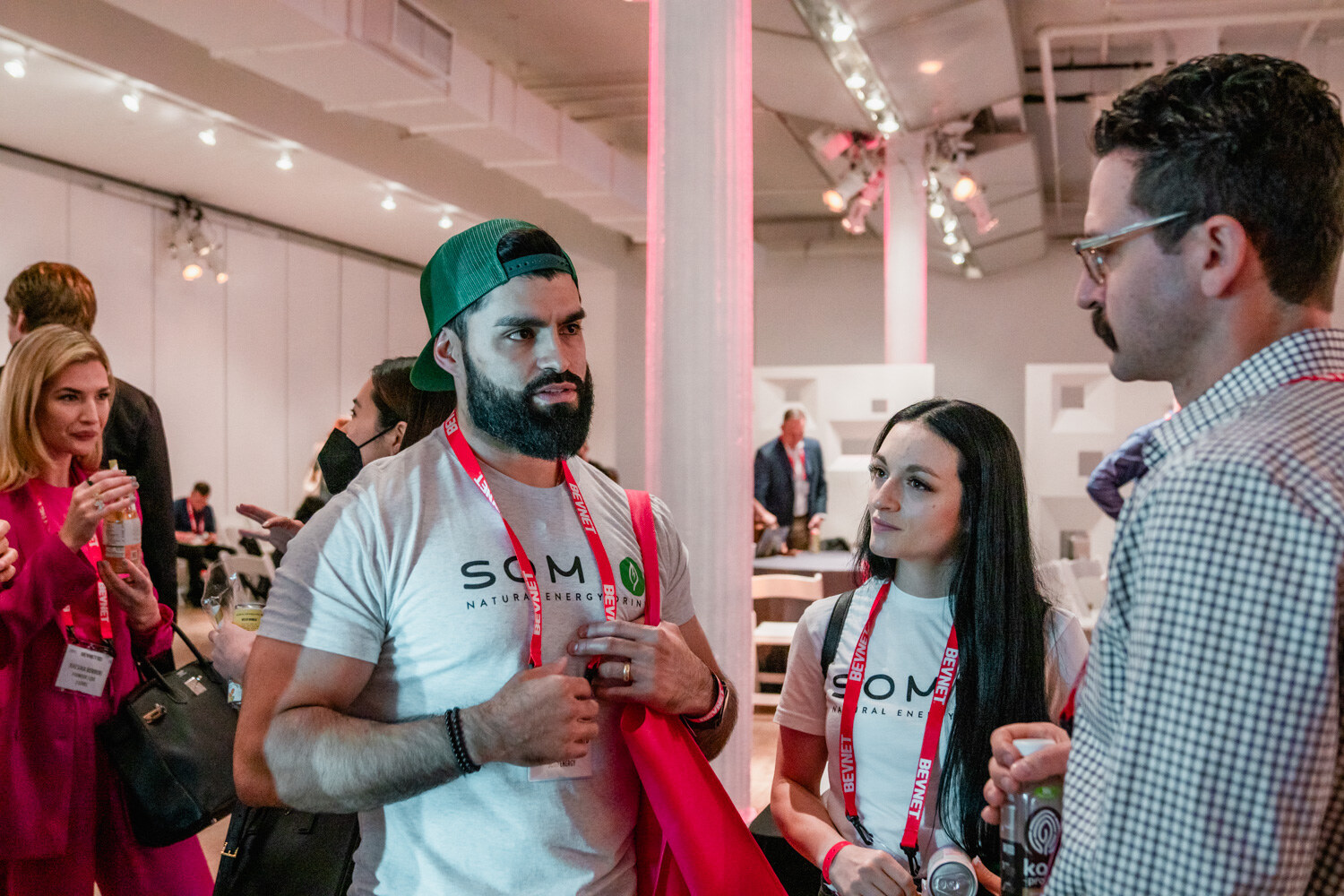 A bearded man in a green cap and white Somi shirt stands near a woman in a Somi shirt and a man in a checked shirt.