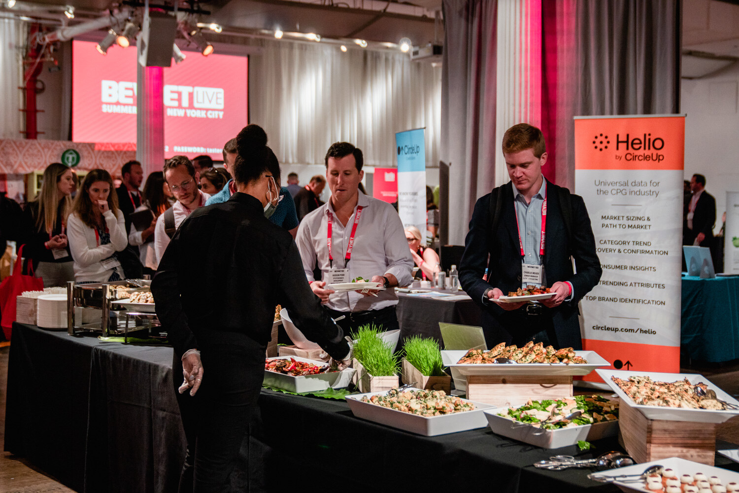 Two men in lanyards pick food from trays on a black-draped buffet table next to an orange Helio by CircleUp banner.