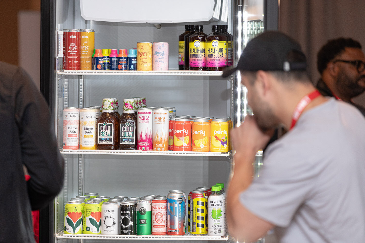 A glass-door cooler of canned drinks including Cold Brew and kombucha, with attendees in lanyards beside it.
