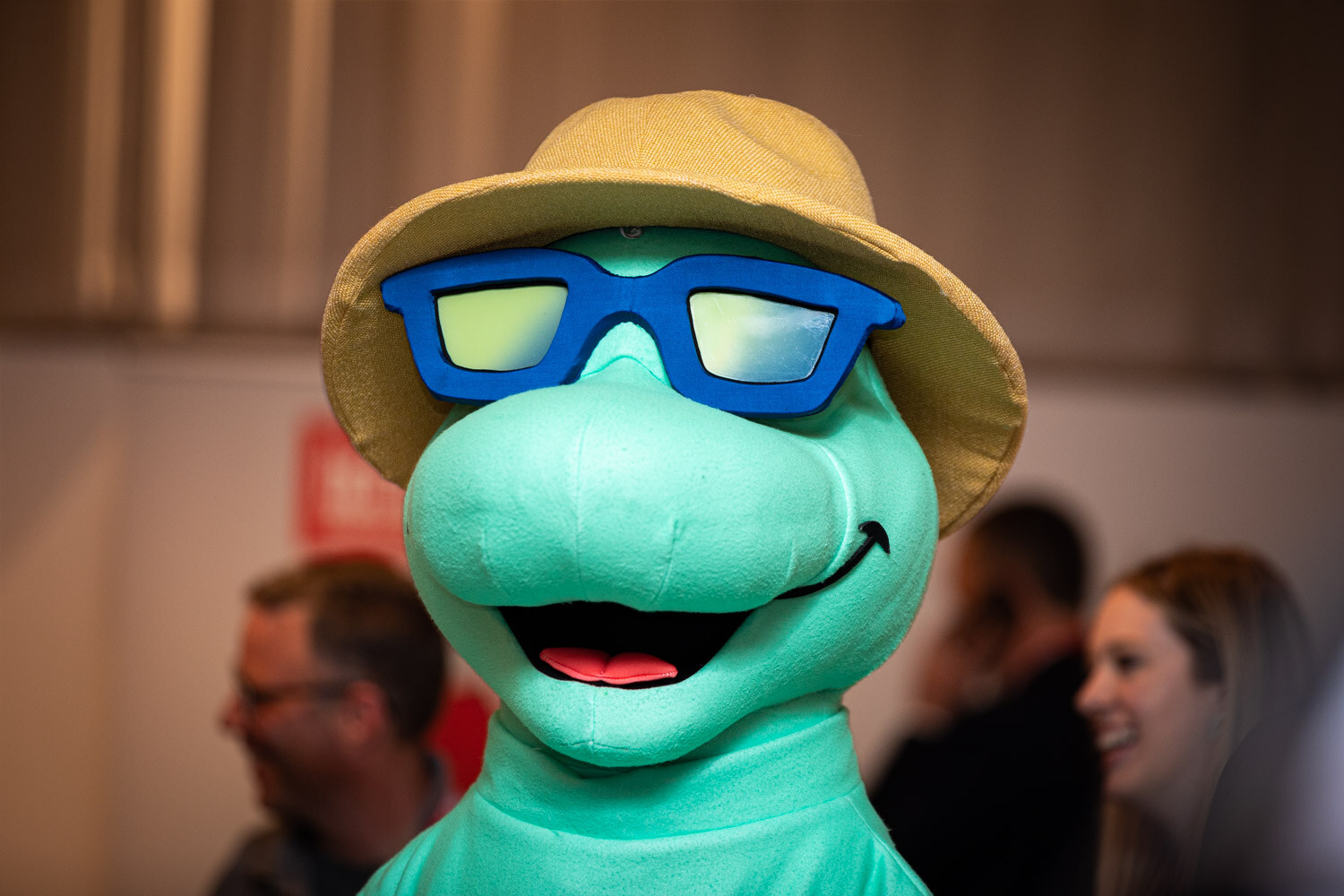 A teal turtle mascot with blue sunglasses and a tan hat in close-up, with out-of-focus attendees behind it.
