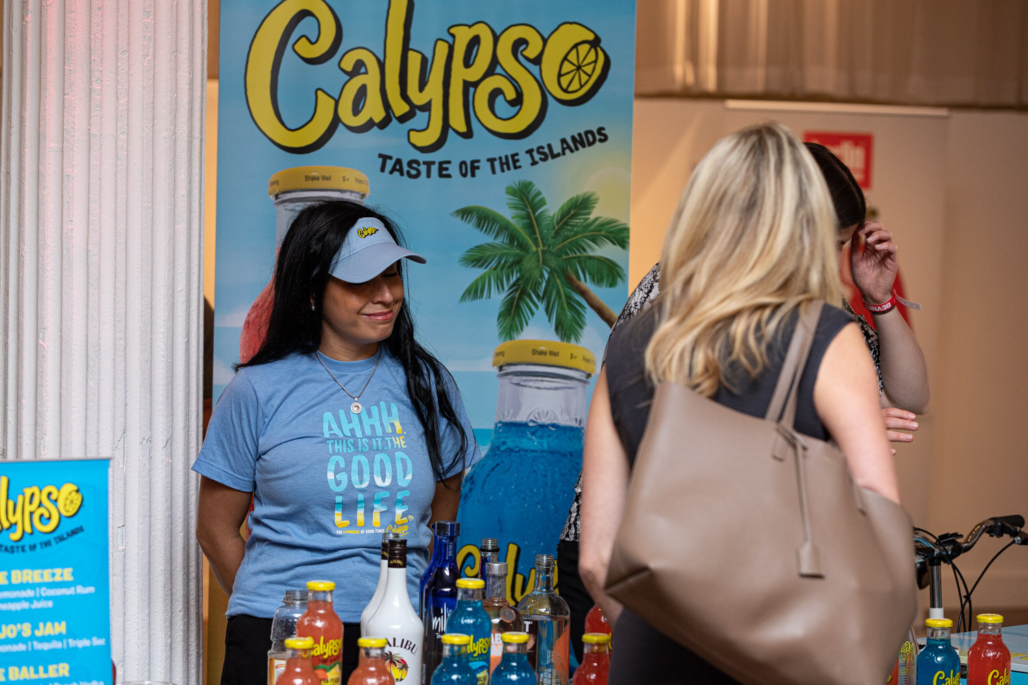 A woman in a blue Good Life t-shirt and a visor stands in front of a Calypso Taste of the Islands banner with bottles on the table.