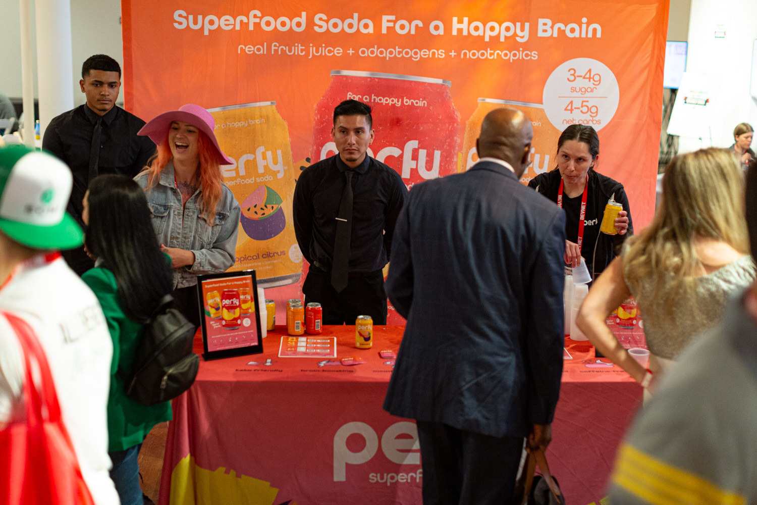 A group of attendees stand in front of an orange Perfy superfood soda banner and a table with an orange tablecloth.