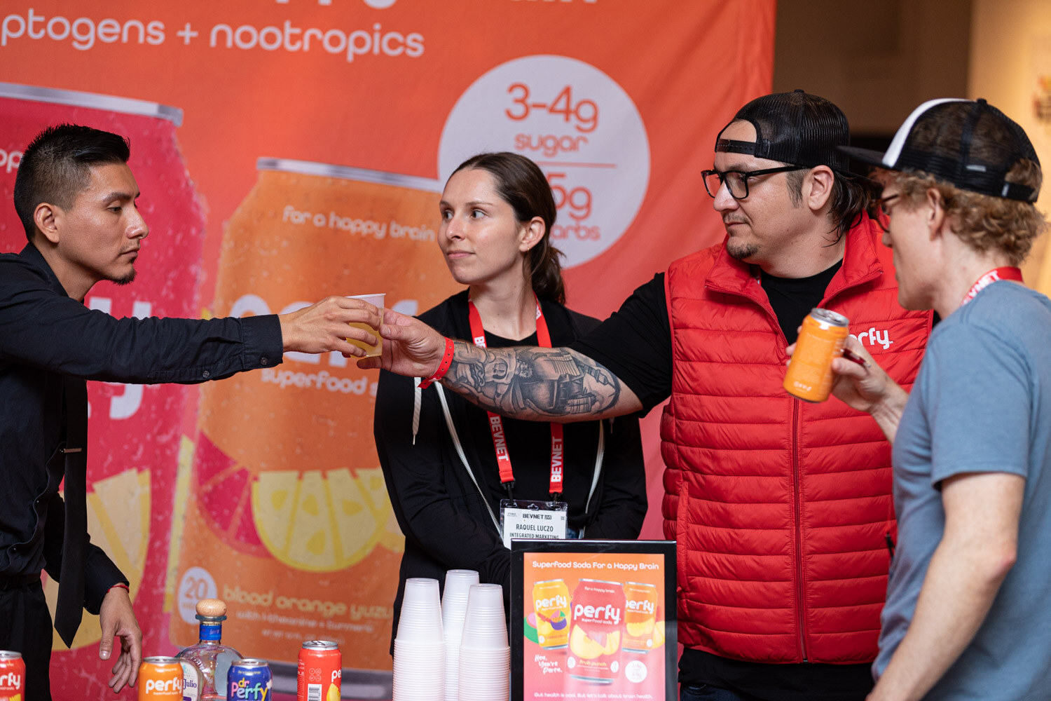 Several people pass cups and cans at a booth with orange Perfy superfood soda banners.