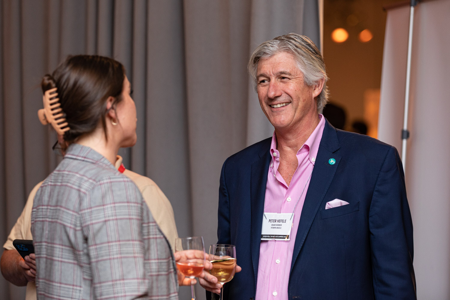 A smiling man in a navy blazer and pink shirt speaks with a woman holding a glass of wine in front of a gray curtain.