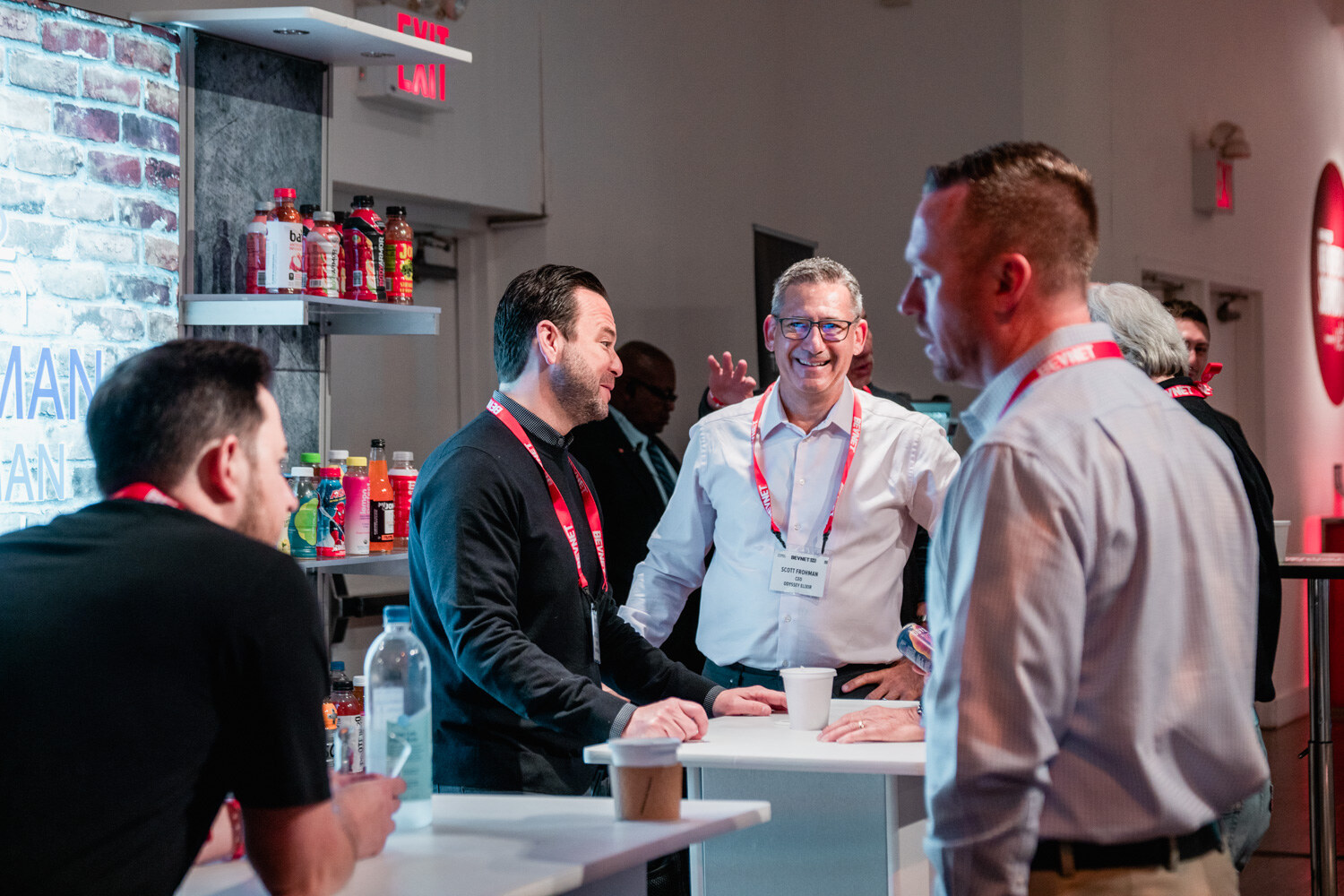Four men in lanyards talk around a white high-top table in front of a brick wall with drinks on shelves.