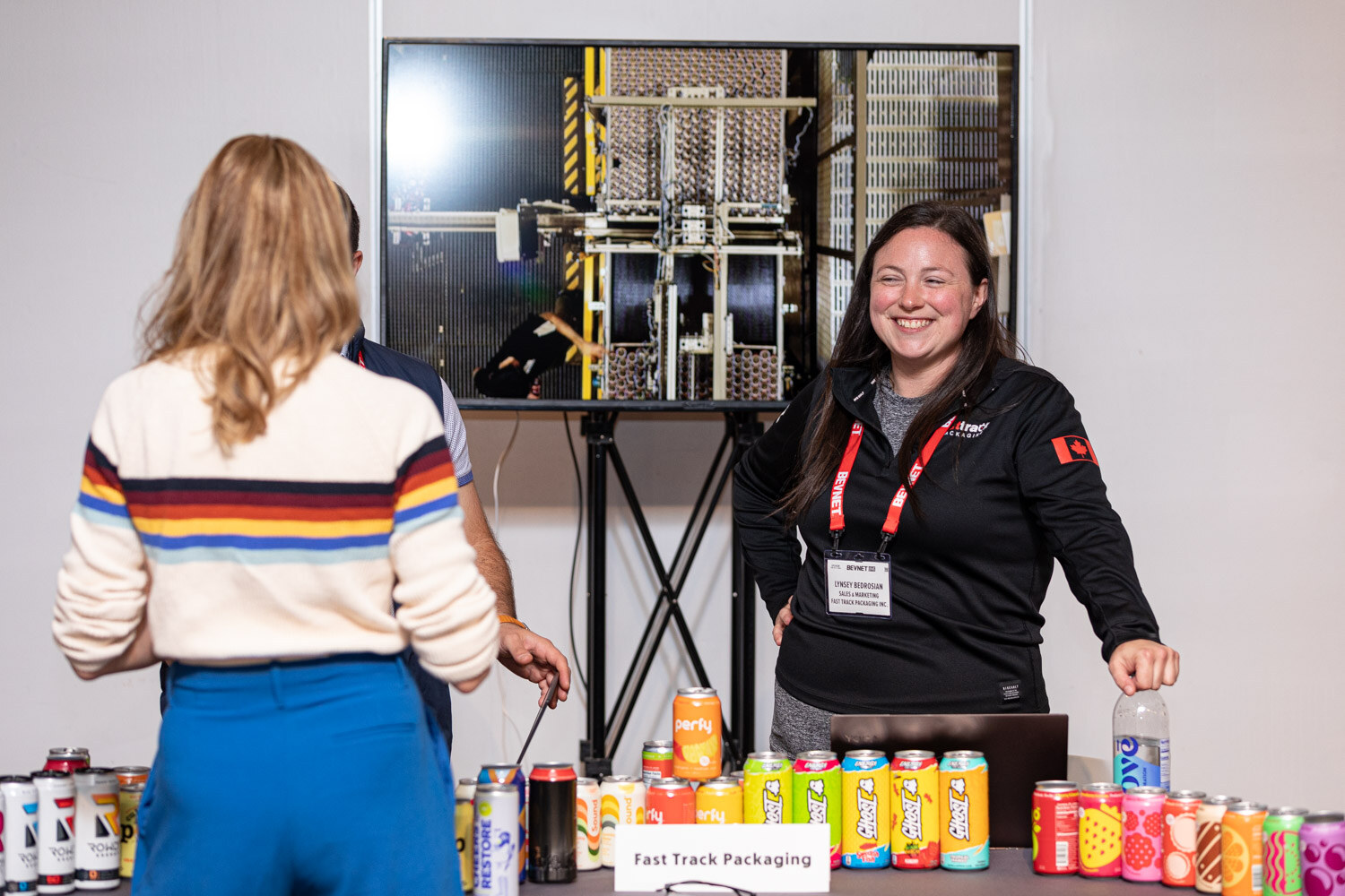 A woman in a black jacket and lanyard smiles behind a table lined with colorful cans, a screen showing machinery above her.