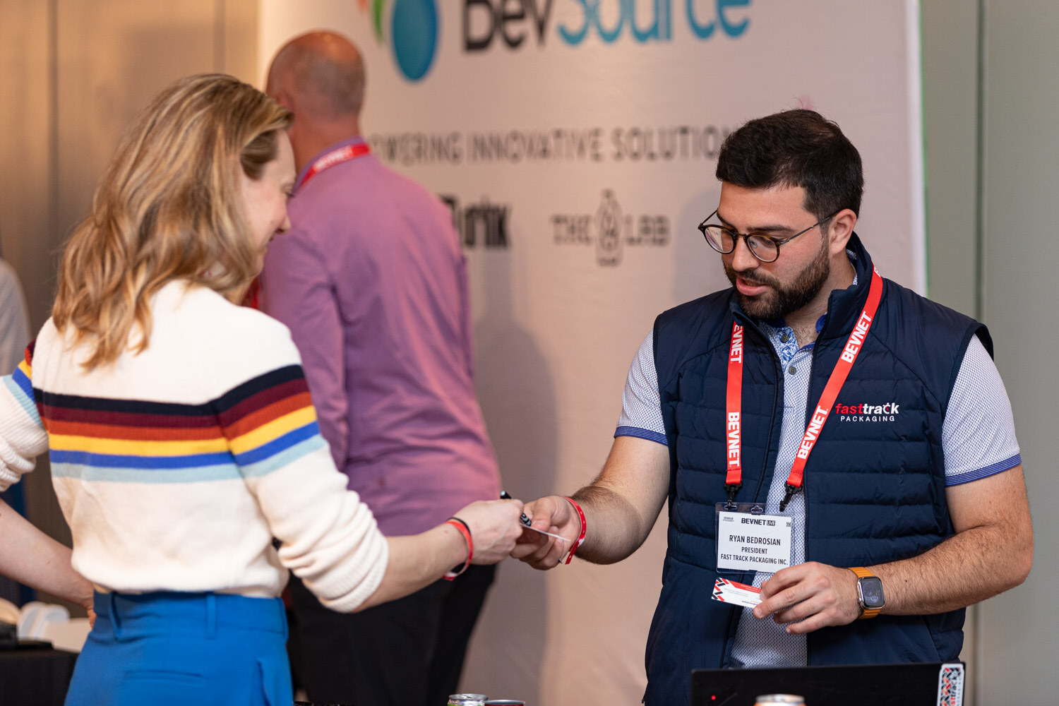 A bearded man in glasses and a navy Fasttrack Packaging vest exchanges a business card with a woman in a striped sweater.
