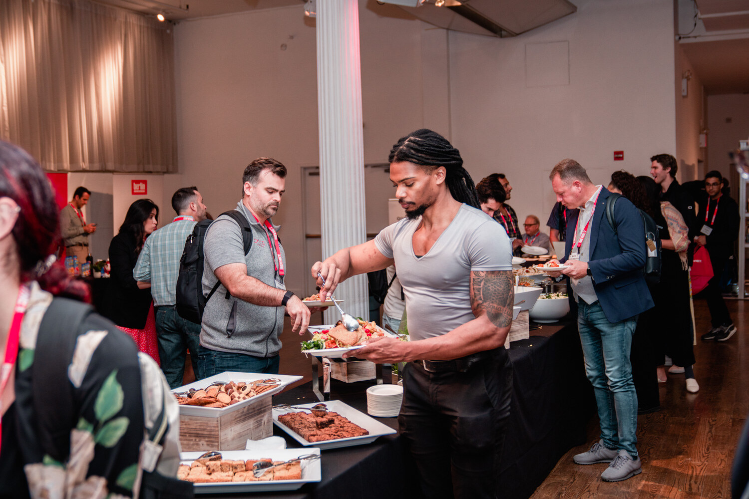 A tattooed man in a gray t-shirt serves himself from a tray at the lunch buffet, with a line of attendees behind him.