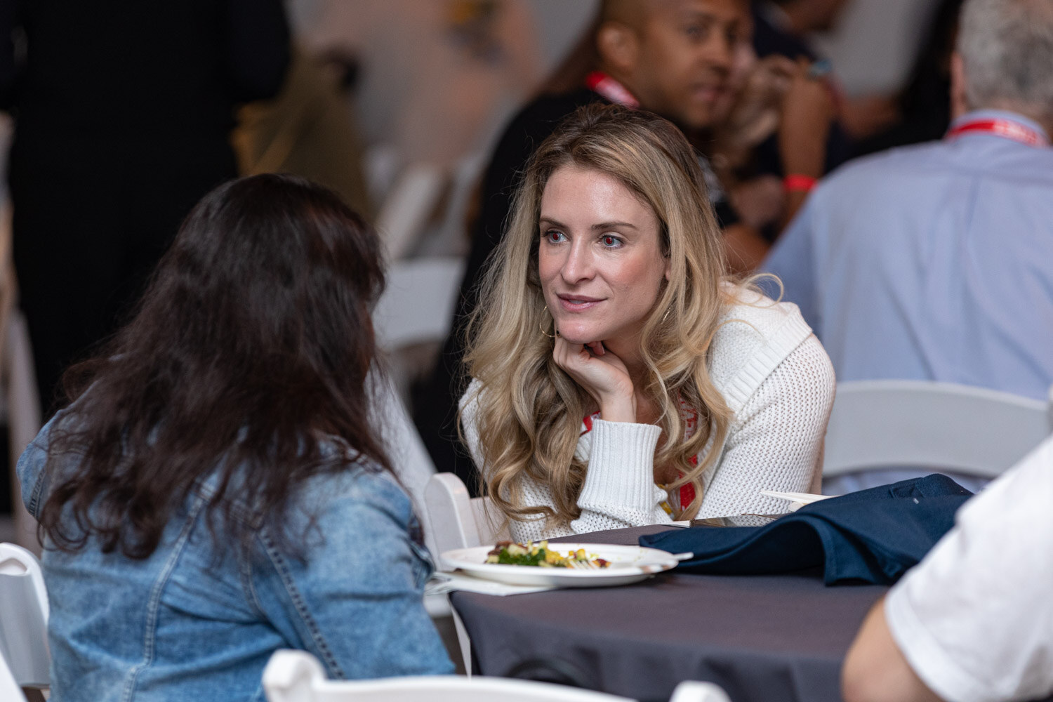 A woman with long blonde hair rests her chin on her hand listening to a woman in a denim jacket, with plates on the table.