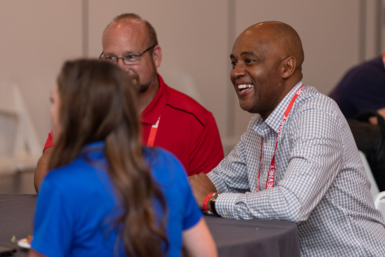 A smiling man in a checked shirt talks with a woman in a blue top and a man in a red polo seated around a table.