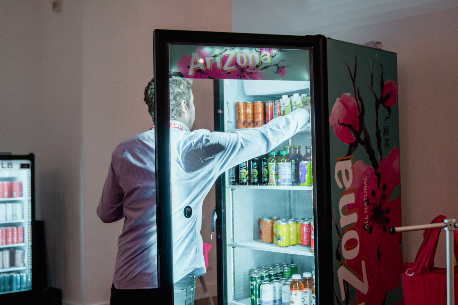 A man in a white shirt reaches into a glowing Arizona-branded cooler stocked with cans and bottles.