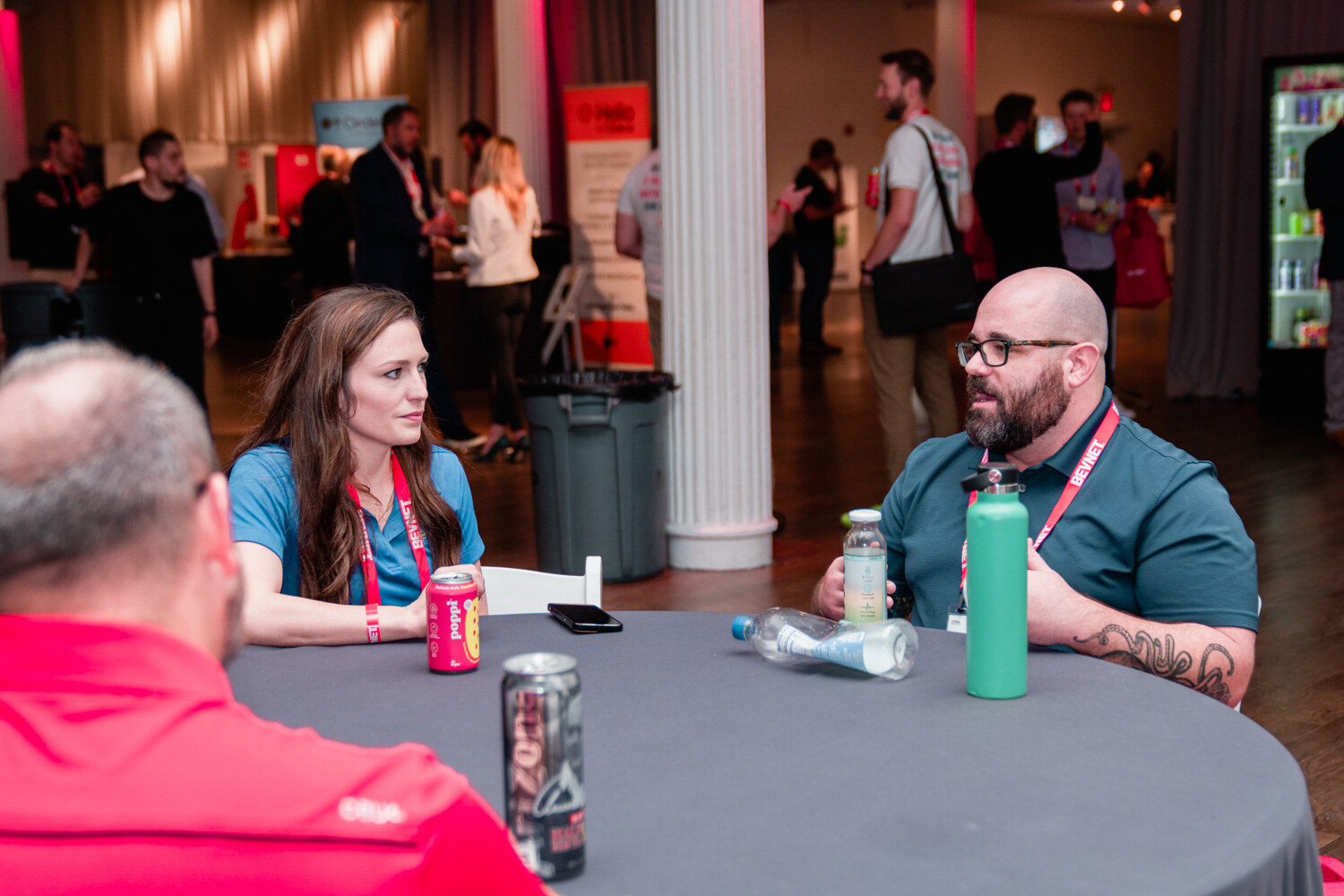 A woman in a teal top and a bearded man with glasses sit at a round table with a Zoa can and a green bottle.