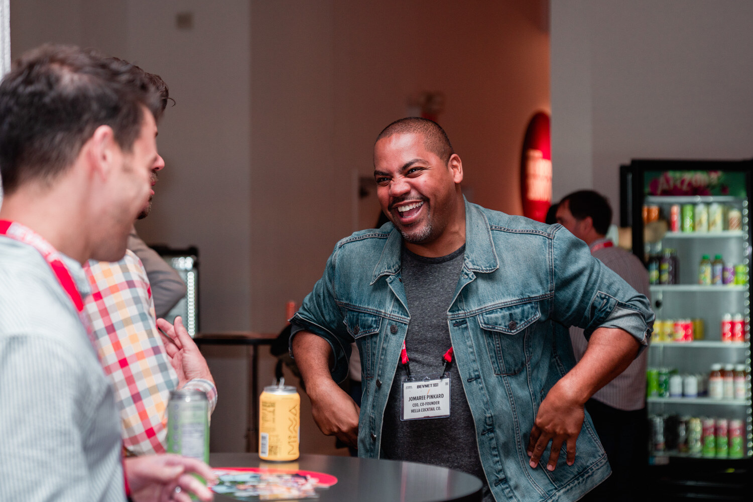 A man in a denim jacket and gray shirt laughs with his hands on his hips, with a cooler of drinks in the background.