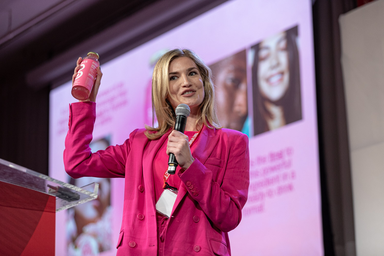 A woman in a hot pink blazer holds a microphone in one hand and a pink bottle in the other, with a pink slide behind her.