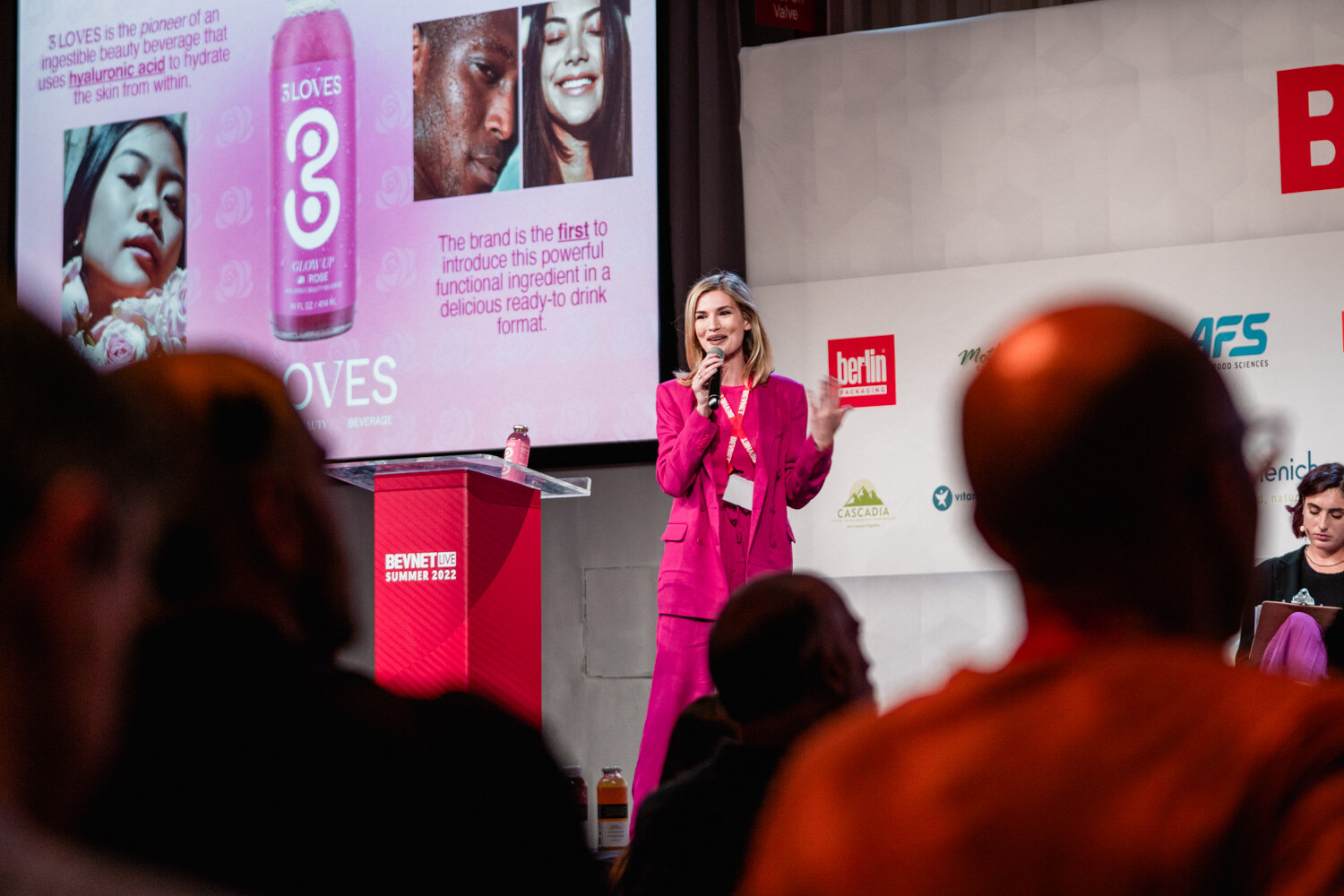 A woman in a hot pink suit stands beside a clear podium with a pink slide of 3LOVES cans behind her.