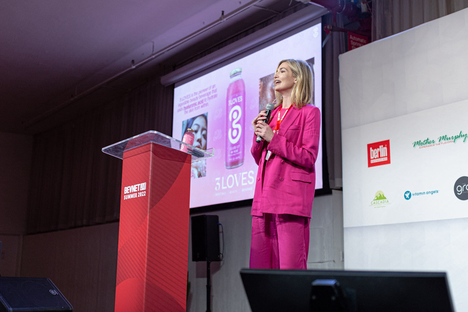 A woman in a pink suit holds a microphone beside a red podium in front of a pink slide with a 3LOVES bottle.