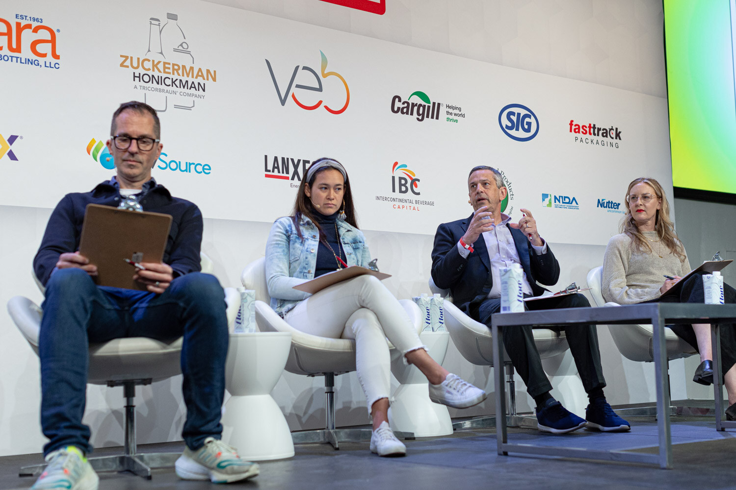 Four people seated in white chairs across a stage with clipboards, a low table in front of them and sponsor logos behind.