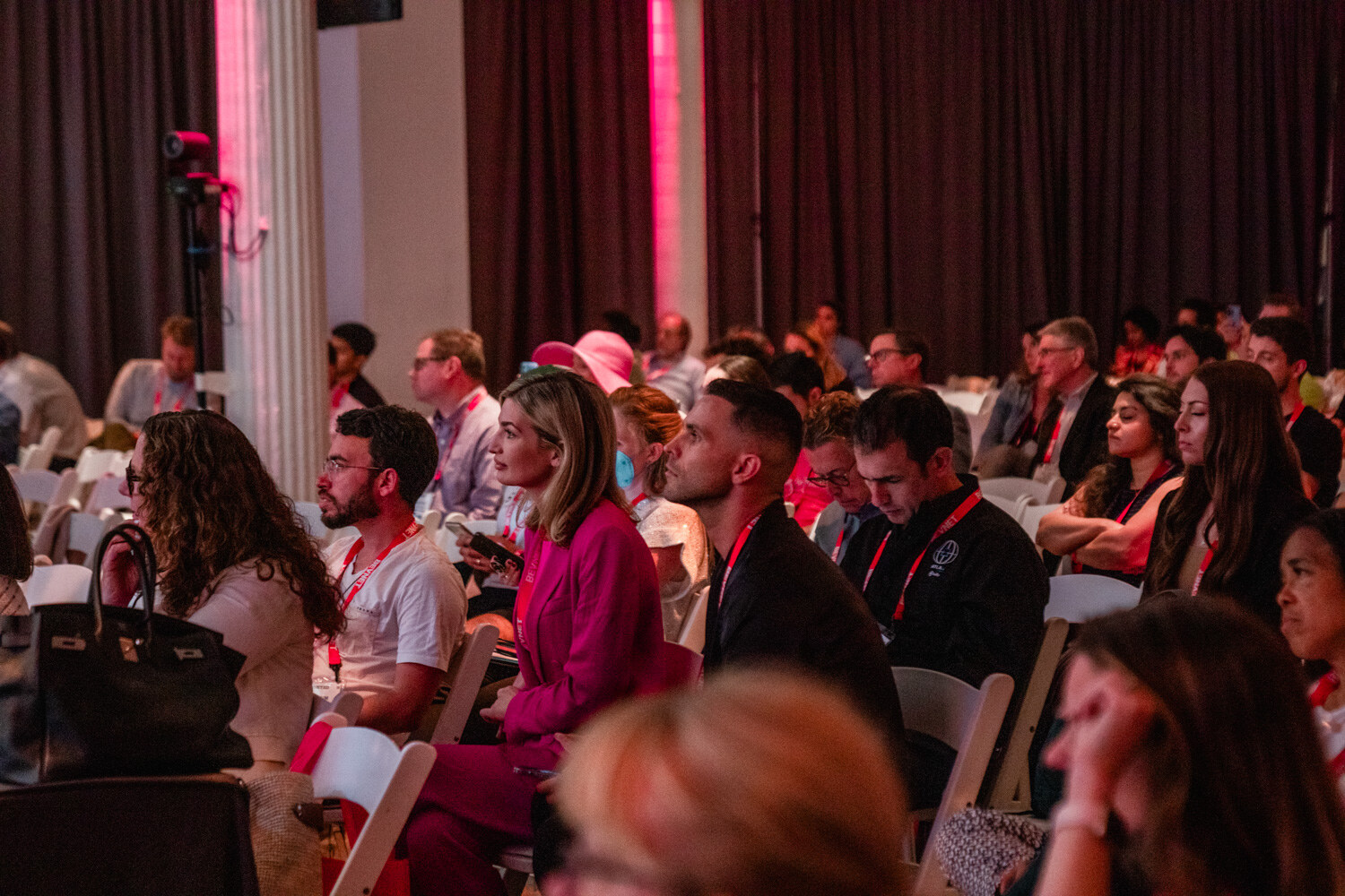 Seated audience members in lanyards watch the stage, a woman in a pink suit in the front row.