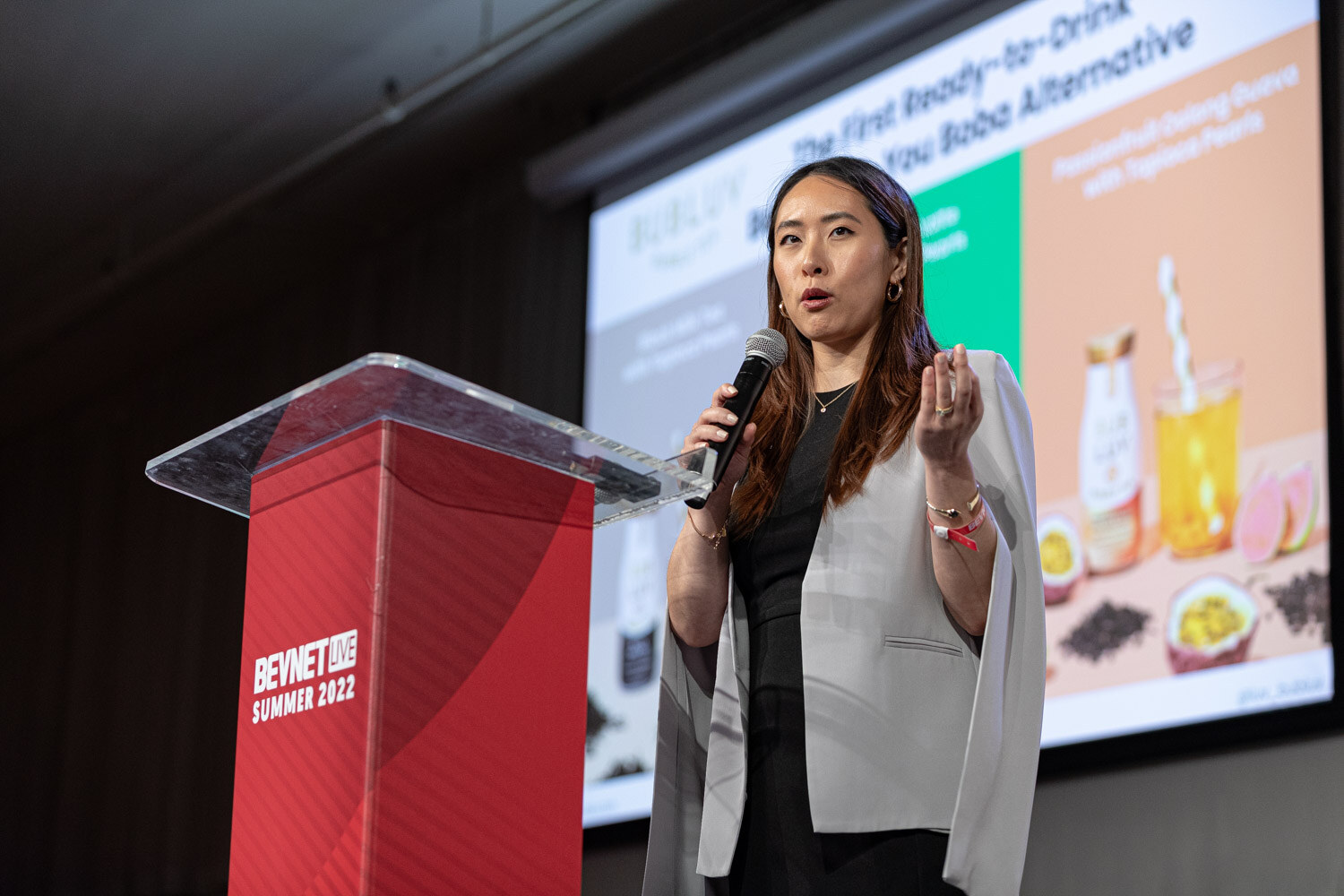 A woman in a gray cape blazer holds a microphone at a red podium in front of a slide reading The First Ready-to-Drink Better-for-You Boba Alternative.