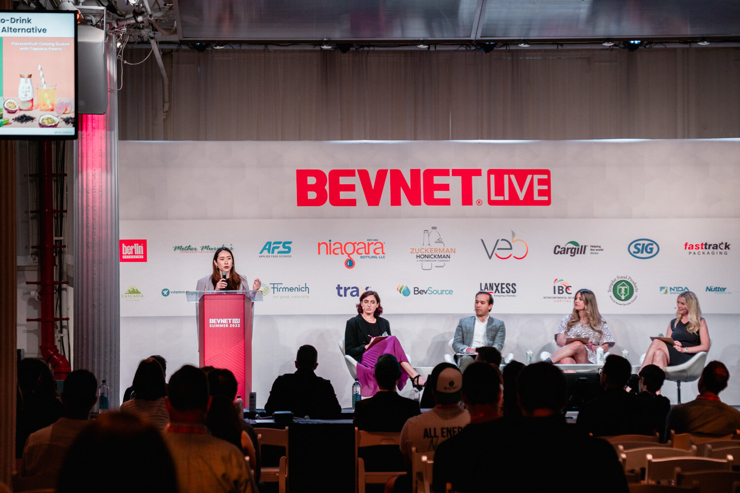 Wide view of a woman in a gray blazer at the podium and four judges seated on stage below the BevNET Live sign.