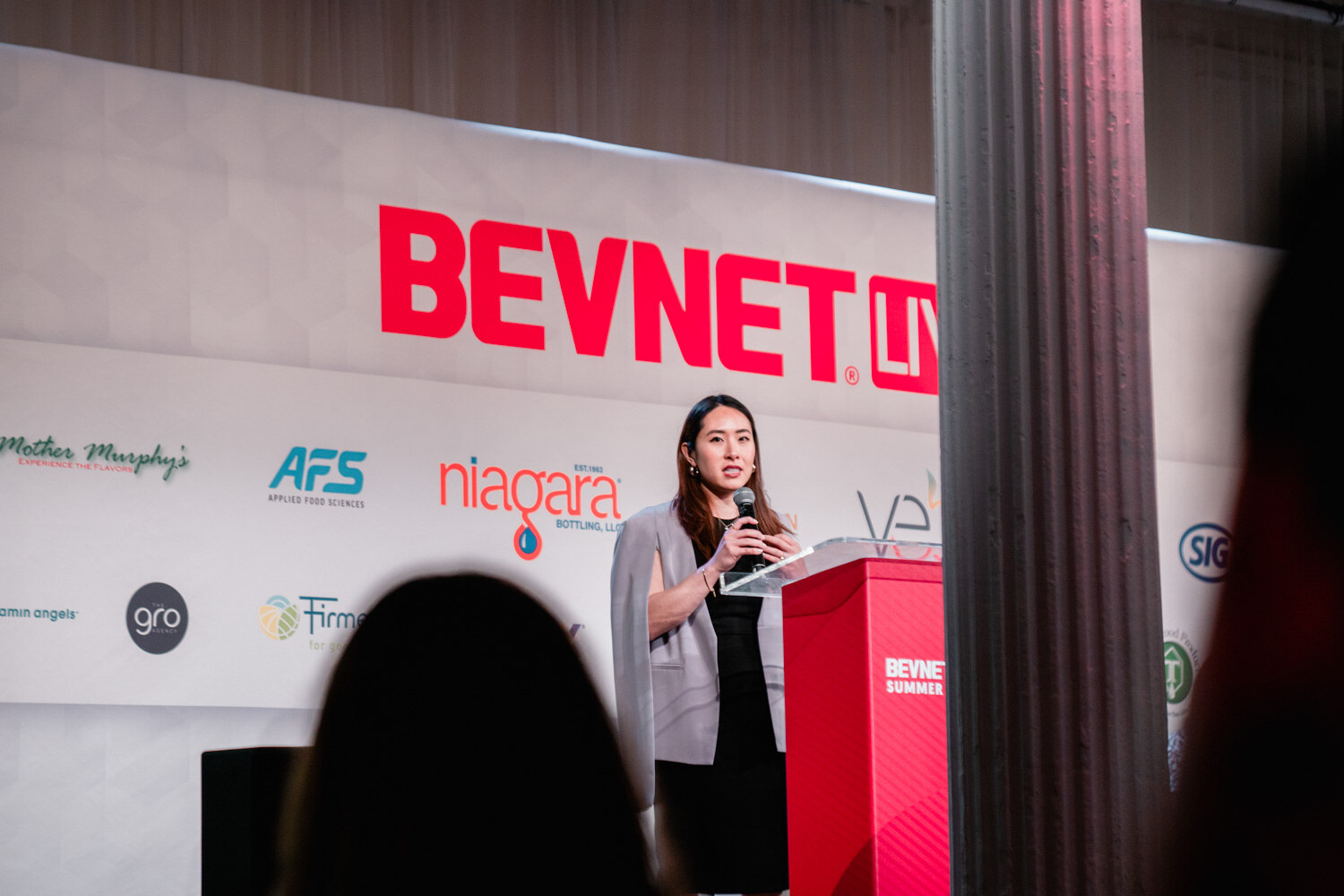 A woman in a gray blazer holds a microphone at a red podium with the BevNET Live logo behind her, a column in the foreground.