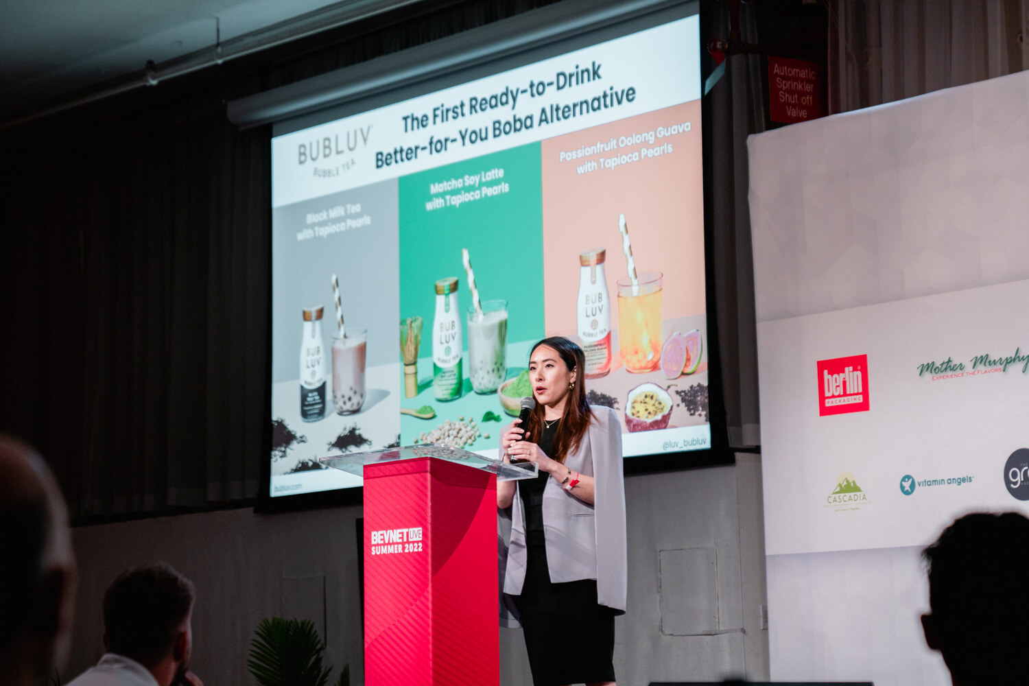 A woman in a gray blazer holds a microphone at a podium while a slide with three bottled drinks and the heading The First Ready-to-Drink Boba shows.