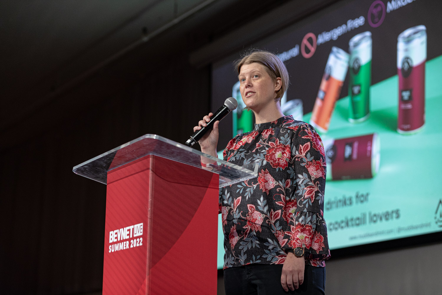 A woman in a floral blouse speaks into a microphone at a red podium, with a screen showing red, green and orange cans behind her.