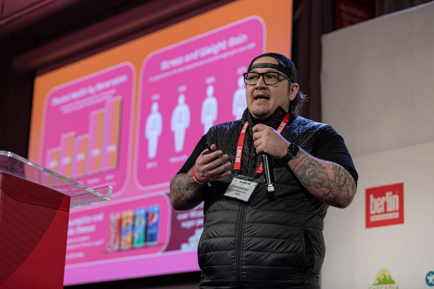 A man in a black vest, backwards cap and glasses holds a microphone and gestures in front of a pink and orange slide with bar charts.