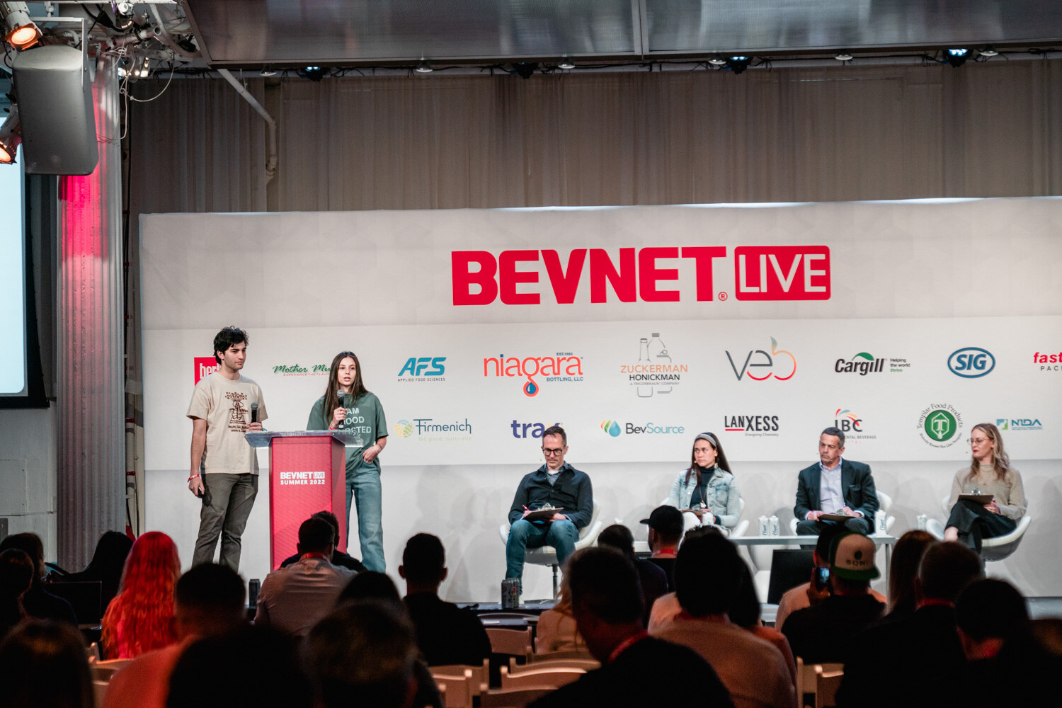 Wide view of two presenters standing at a red podium on a stage under a BevNET Live sign, with four seated judges to their right and the audience in the foreground.