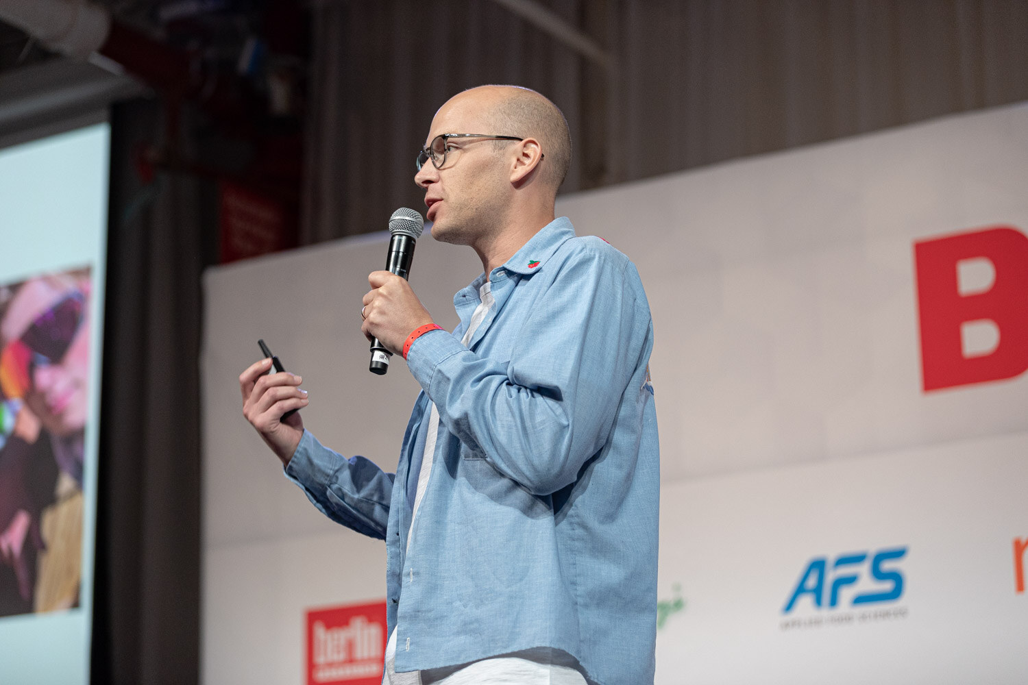 A bald man with glasses in a blue shirt holds a microphone and a clicker as he speaks on stage.