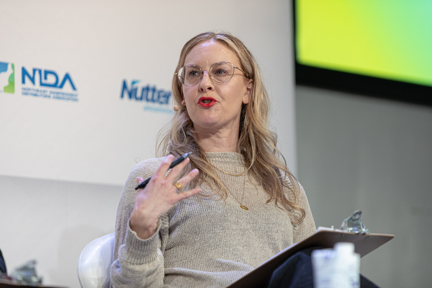 A woman with long blonde hair and round glasses holds a pen and gestures while speaking, with a clipboard on her lap.