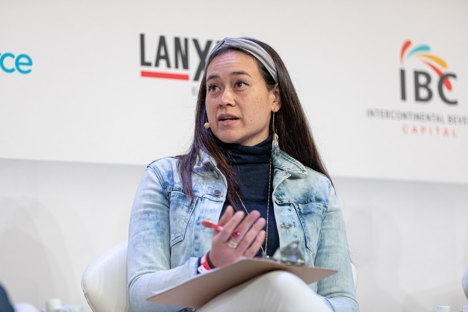 A woman in a denim jacket and head scarf glances to her side with a pen and clipboard, in front of IBC and Lanxess signs.