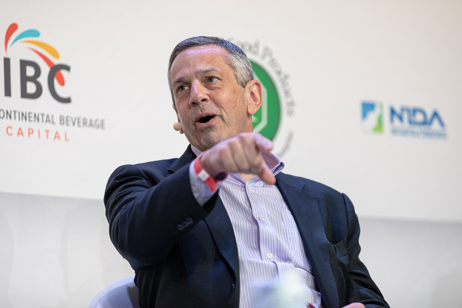 A man in a navy blazer and striped shirt points his finger forward while speaking, with IBC and NIDA logos behind him.
