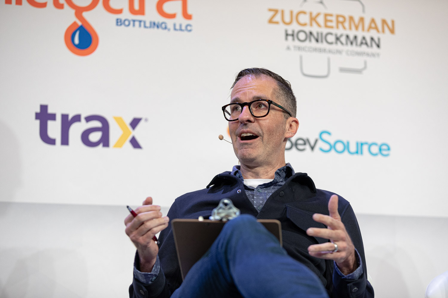 A man in a dark jacket and black glasses gestures with both hands, with Trax and BevSource signs behind him.