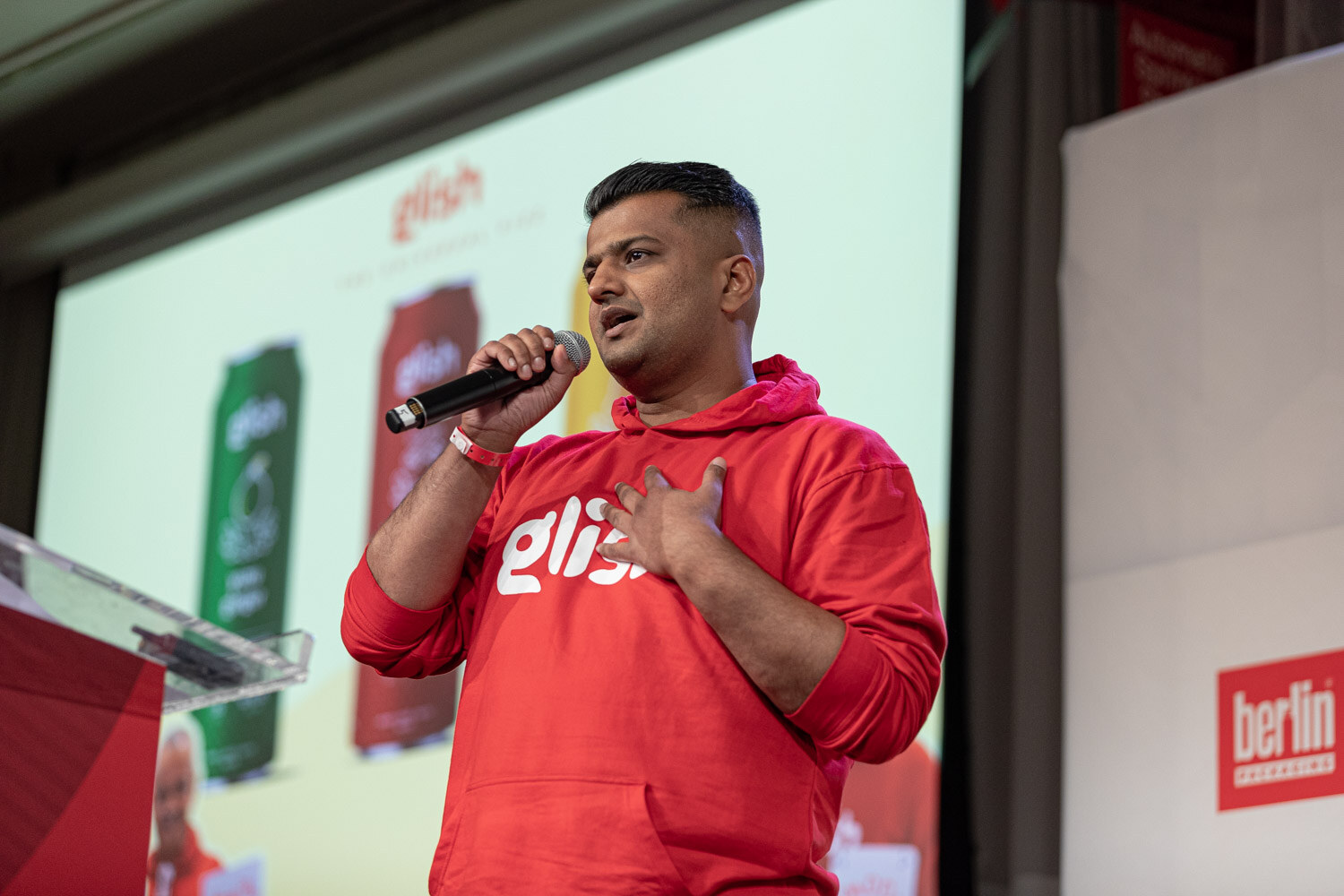 A man in a red hooded sweatshirt with Glish printed on it holds a microphone in front of a slide showing green and red cans.