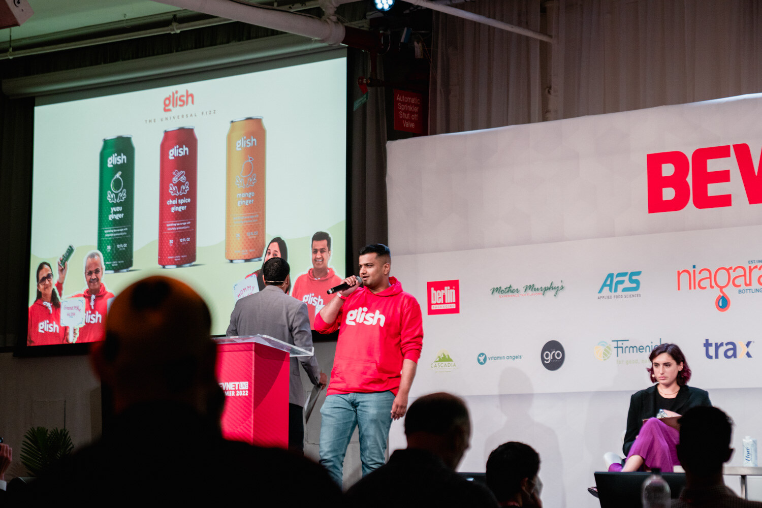A man in a red Glish hoodie holds a microphone at a podium in front of a screen of green, red and orange Glish cans.