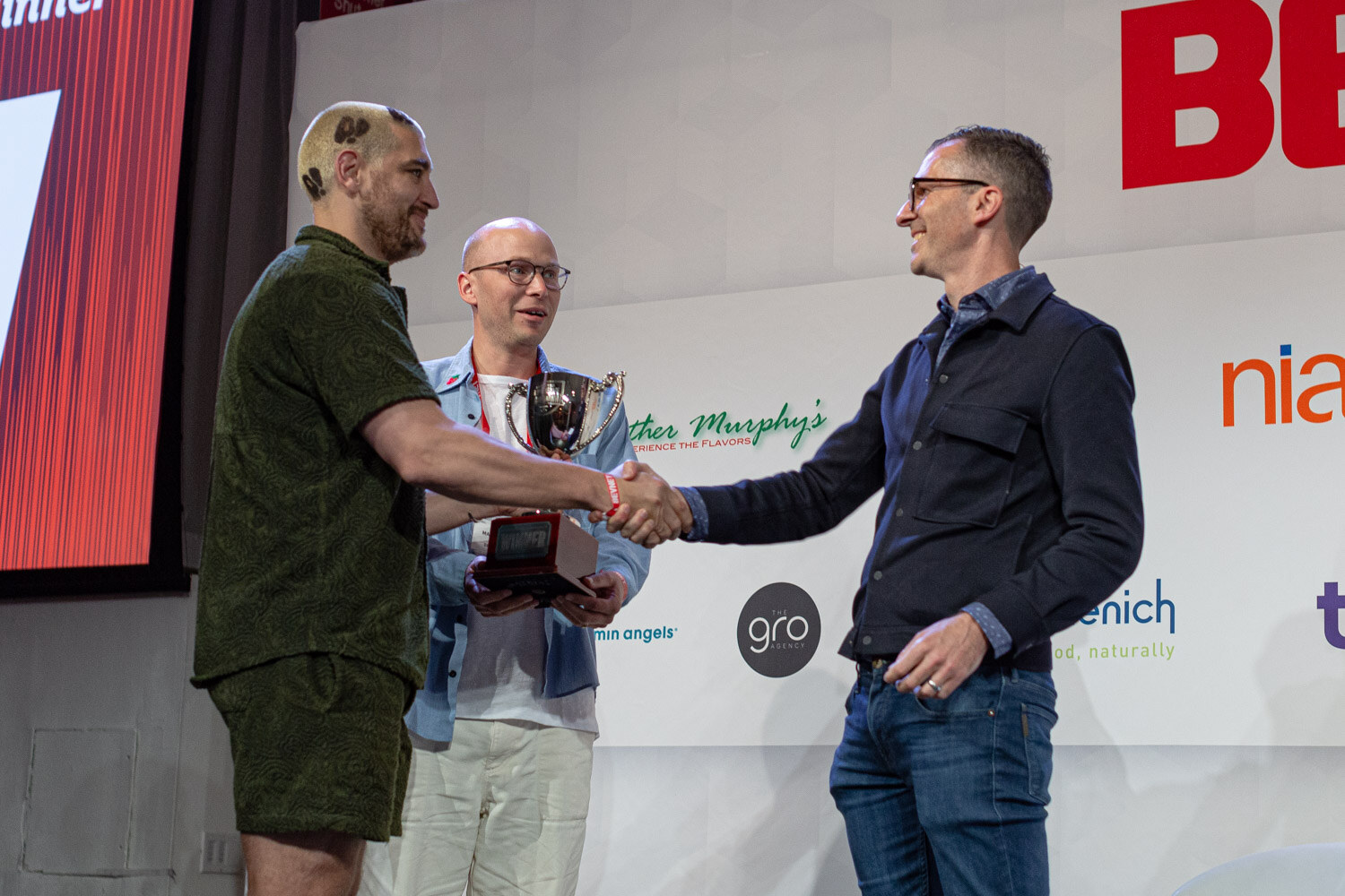A man in a green shirt shakes hands with a man in a navy jacket as a bald man in a blue shirt holds a small trophy between them.