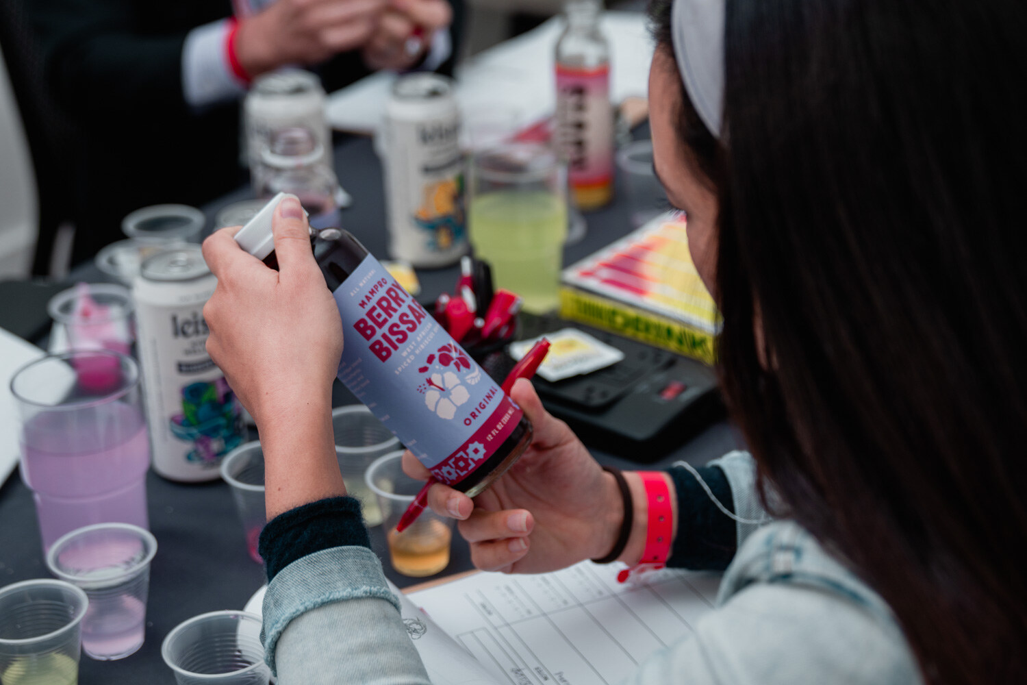 A woman in a denim jacket holds a bottle labeled Berry Bissap beside cups and cans of Leisure drinks on a black tablecloth.