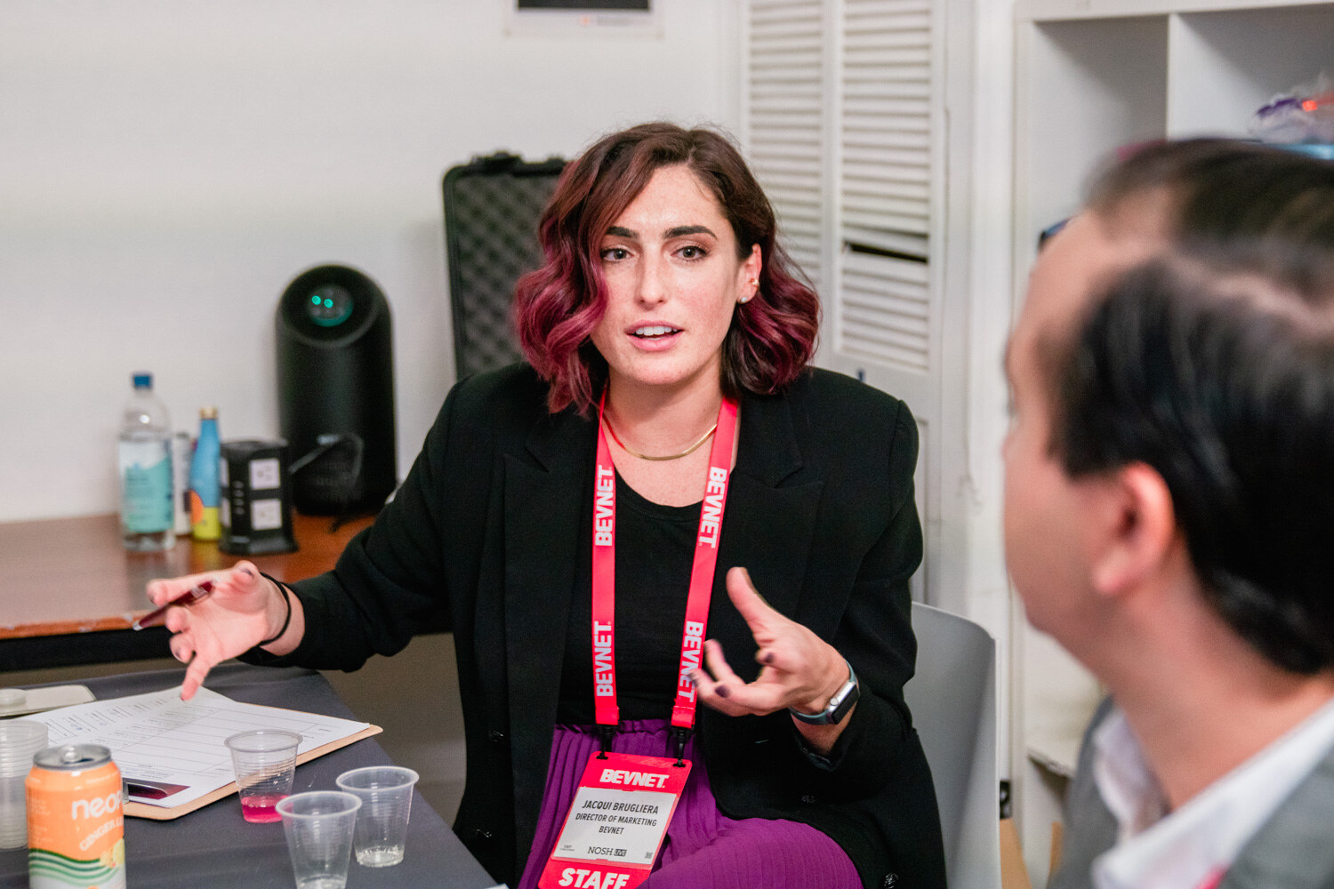 A woman with dark red hair in a black blazer and a BevNET staff lanyard gestures while talking, with sample cups and a clipboard on the table.