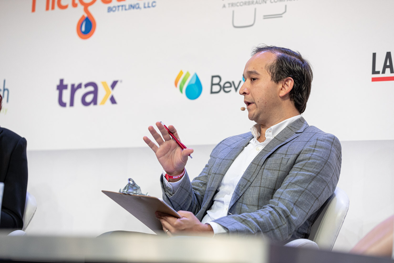 A man in a gray plaid blazer gestures with a pen in his right hand, a clipboard on his lap, Trax and BevSource logos behind him.