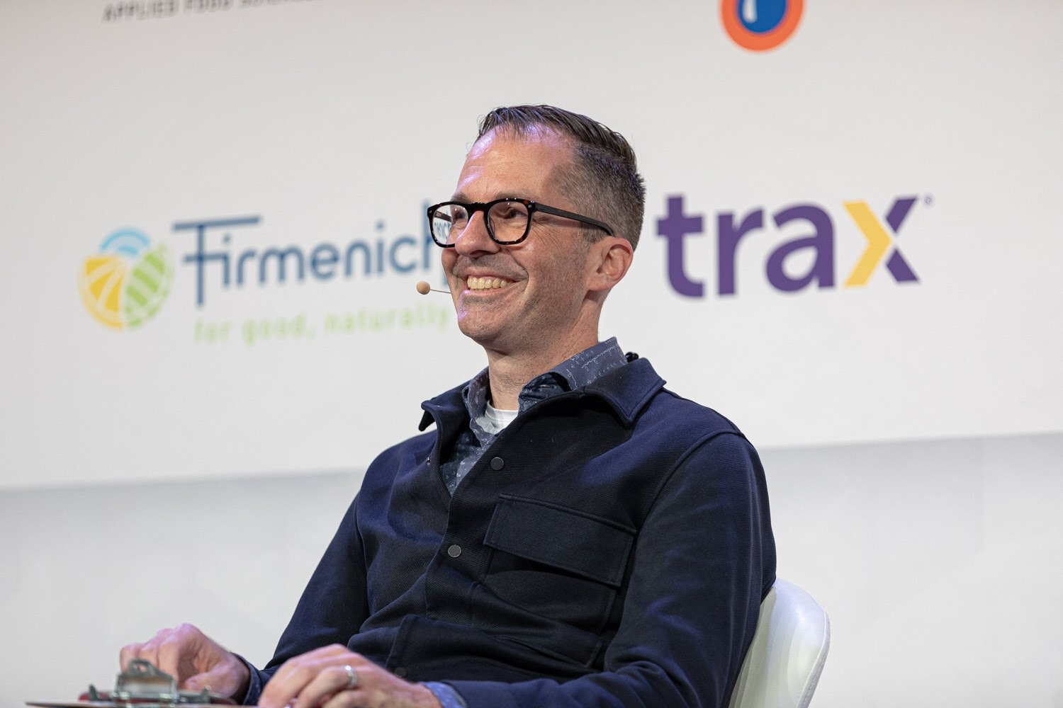 A man in glasses and a navy jacket smiles with a clipboard on his lap in front of Firmenich and Trax signs.
