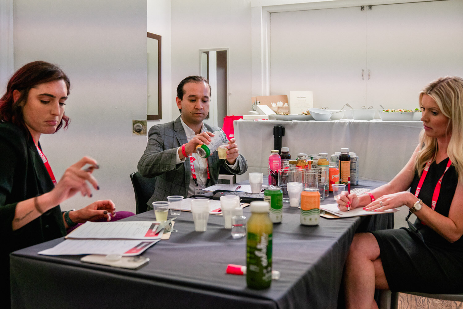 Three people in lanyards sit at a table covered with sample cups and drinks, tasting and writing on score sheets.