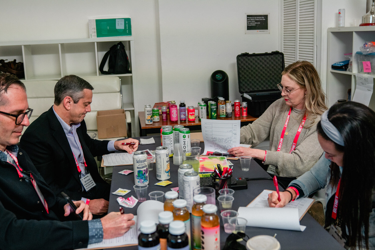 Four people in lanyards sit around a long table covered with cans, bottles and cups, filling out scoresheets.