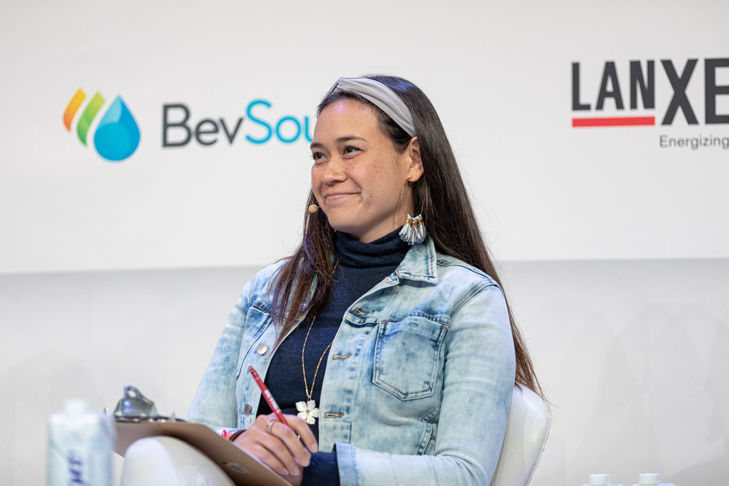 A woman in a denim jacket and head scarf smiles with a clipboard and red pen in front of BevSource and Lanxess signs.