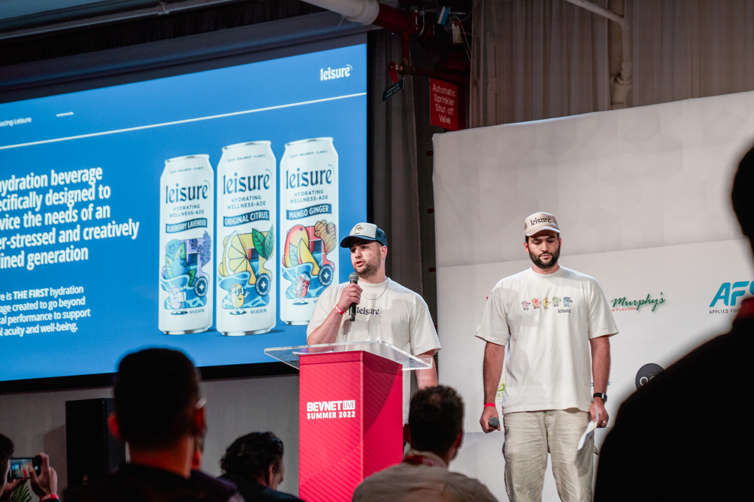 Two men in cream shirts and caps stand at a red podium in front of a blue slide showing three Leisure cans.