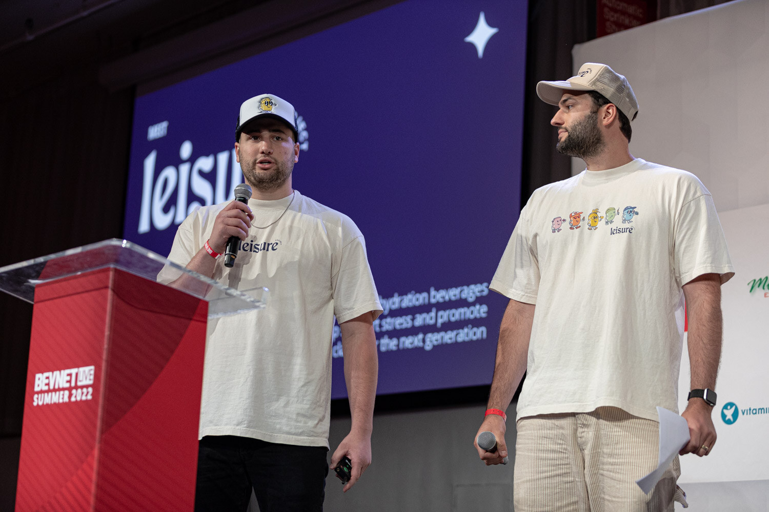 Two men in cream Leisure t-shirts and caps, one holding a microphone, stand beside a red podium in front of a blue Leisure slide.