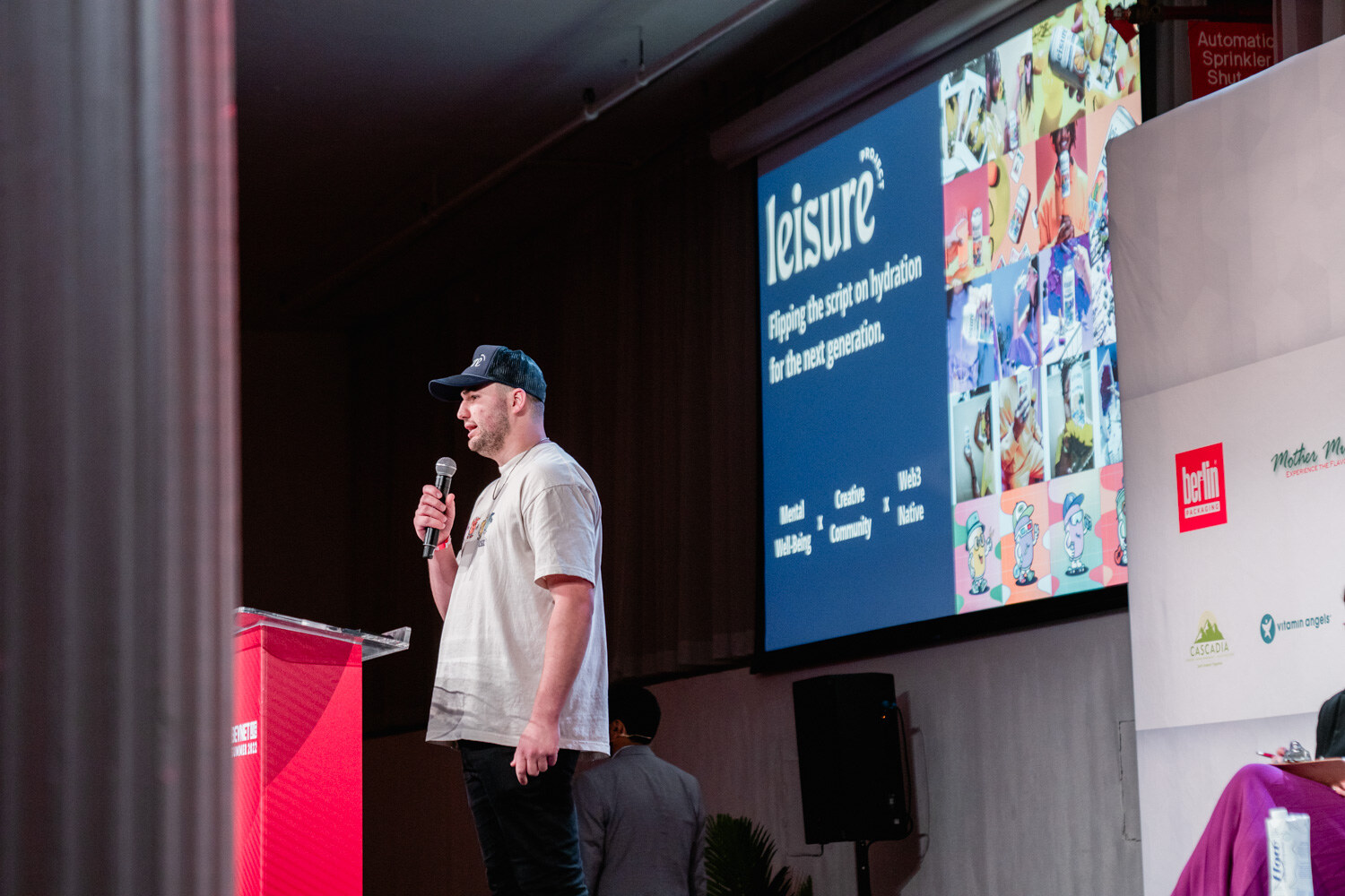 A man in a cream t-shirt and dark cap stands beside a podium as a blue Leisure slide with a collage of images shows behind him.
