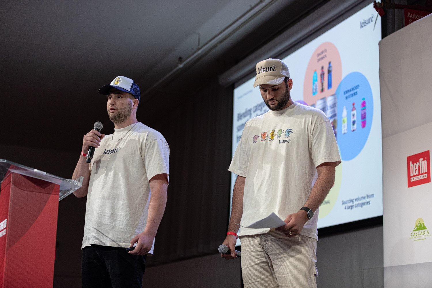Two men in cream t-shirts and caps stand at the podium, one speaking into a microphone while the other looks down at his papers.